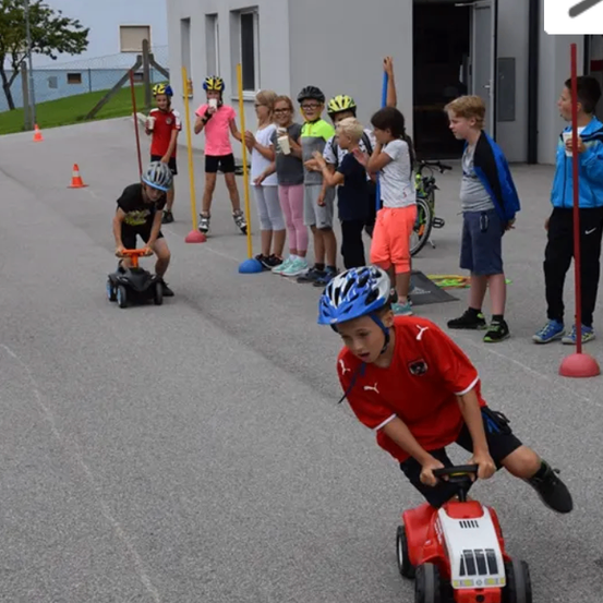 Eine Gruppe von Kindern in Helmen fährt mit Spielzeugautos auf einem Parkplatz. Einige von ihnen beobachten, und es gibt Verkehrskegel und Stangen im Bereich.