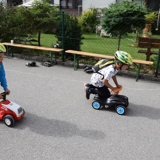 Zwei Kinder fahren mit Spielzeugautos auf der Straße. Sie tragen Helme und Turnschuhe. Im Hintergrund befinden sich Holzbänke und Topfpflanzen.