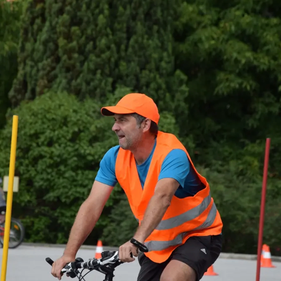Ein Mann fährt auf einer von Bäumen gesäumten Straße mit einem Fahrrad, trägt eine orange Weste, ein blaues Hemd und eine Kappe.