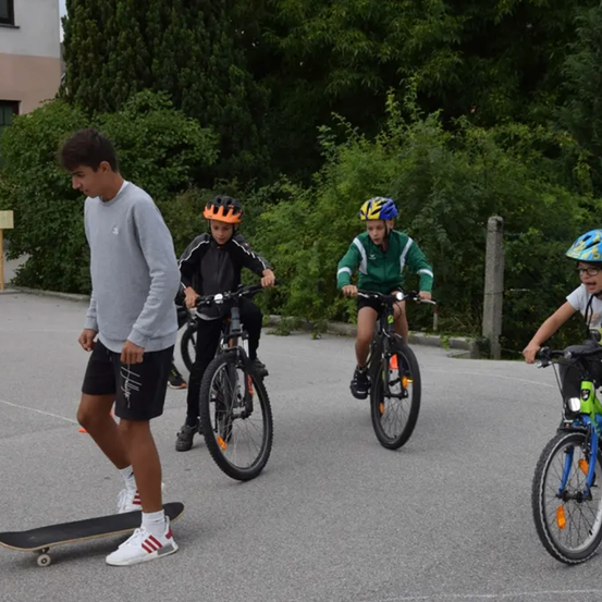Eine Gruppe von Jungen fährt auf einem asphaltierten Bereich mit Fahrrädern. Ein Junge trägt einen Helm und ein anderer hält ein Skateboard. Im Hintergrund befinden sich Bäume und Pflanzen.