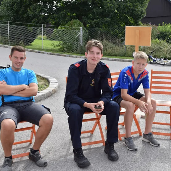 Drei Jungen sitzen auf orangefarbenen Stühlen in einem Außenbereich. Der Junge auf der linken Seite trägt ein blaues T-Shirt und schwarze Shorts. Der Junge in der Mitte trägt eine Uniform mit einem roten Kreuz. Der Junge auf der rechten Seite trägt ein blaues T-Shirt mit rotem Streifen. Ein Fahrrad ist hinter ihnen geparkt.
