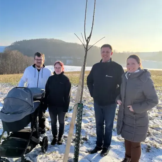 Vier Personen stehen im Schnee mit einem Kinderwagen und einem jungen Baum im Vordergrund.