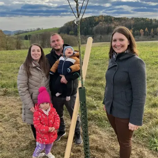 Eine vierköpfige Familie steht in einem Feld neben einem jungen Baum und lächelt in die Kamera. Der Mann hält ein Baby und die Frau neben ihm ein Kleinkind. Die Frau rechts trägt einen grauen Mantel und der Mann einen schwarzen.