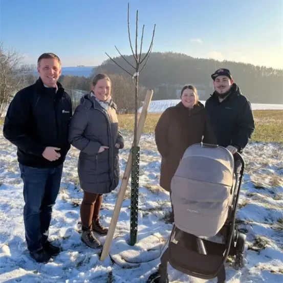 Vier Personen stehen im Schnee neben einem frisch gepflanzten Baum und lächeln für ein Foto. Ein Kinderwagen steht neben ihnen.