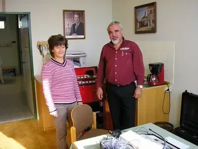 Zwei Personen stehen in einem Zimmer mit Möbeln und Geräten. Ein Mann trägt ein rotes Hemd und eine Uhr, während eine Frau ein gestreiftes Hemd trägt. Ein Bilderrahmen hängt an der Wand.