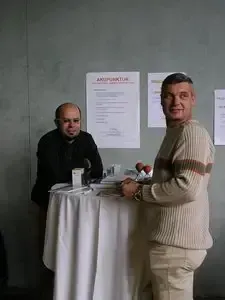 Zwei Männer stehen neben einem Tisch mit einer weißen Tischdecke, einer von ihnen lächelt, und ein Schild mit dem Text 'Ankaufaktur' an der Wand hinter ihnen.