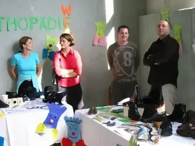 Eine Gruppe von Menschen steht in einem Raum mit Tischen, auf denen verschiedene Gegenstände ausgestellt sind. Der Raum hat eine Wand mit dem Wort 'THOPADiE'. Die Gruppe besteht aus zwei Frauen und zwei Männern, von denen einer eine Brille trägt.