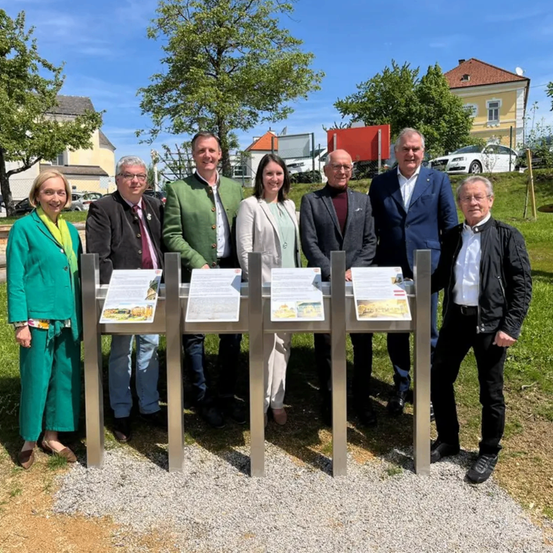Sieben Personen stehen vor einem Denkmal mit Tafeln. Dahinter befinden sich Gebäude und Bäume.