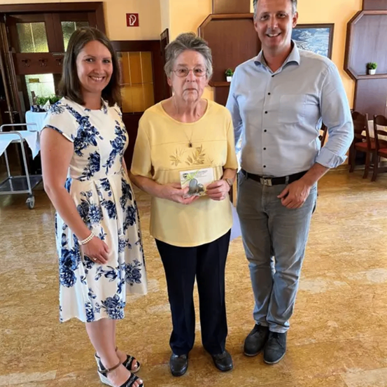 Drei Personen stehen eng beieinander. Eine Frau in einem gelben T-Shirt hält ein Zertifikat in der Hand, und die anderen beiden lächeln. Sie stehen auf einem Holzboden.