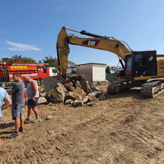 Ein CAT-Bagger entfernt Steine von einer Baustelle, während zwei Männer beobachten, und ein Feuerwehrwagen ist in der Nähe geparkt.
