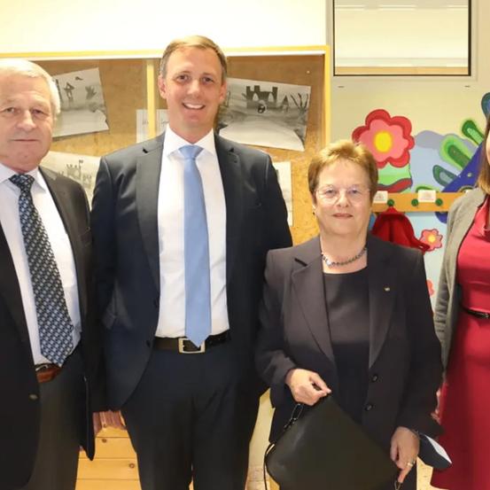 Drei Personen stehen nebeneinander in einem Raum. Sie sind alle in formeller Kleidung gekleidet. Die Frau in der Mitte hält eine schwarze Handtasche. Hinter ihnen befindet sich ein Holzregal mit einigen Papieren und Zeichnungen.