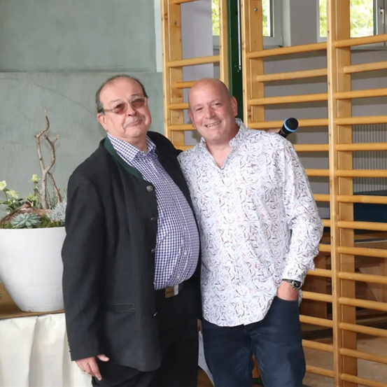 Zwei Männer stehen nebeneinander und lächeln in die Kamera. Sie sind in Anzügen gekleidet und stehen vor einer Holzwand.