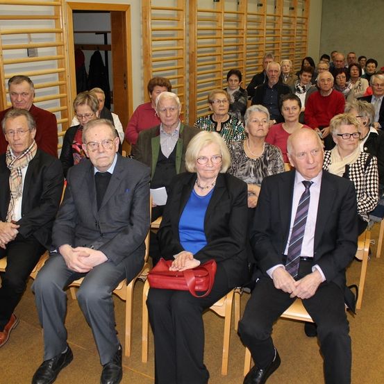 Eine Gruppe von Menschen in formeller Kleidung sitzt auf Holzstühlen in einem Raum, einige tragen Brillen und andere halten rote Handtaschen.