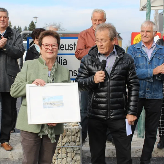 Zwei ältere Männer und zwei ältere Frauen stehen nebeneinander auf einem Bürgersteig. Ein Mann hält ein Mikrofon und spricht, während eine Frau ein gerahmtes Bild hält.