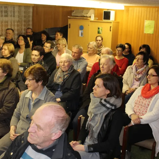 Ein Raum voller Menschen, die auf Stühlen sitzen und einer Präsentation lauschen. Der Raum hat Holzwände und ein gelbes Papier, das an einer der Wände befestigt ist.