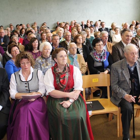 Eine große Gruppe von Menschen, meist älterer Menschen, sitzt in einem Konferenzraum in Stühlen. Einige tragen Brillen und manche halten Bücher. Der Raum hat eine weiße Wand.