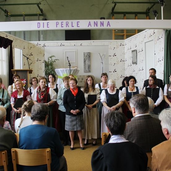Ein Chor von Frauen in traditioneller Kleidung singt ein Lied in einem Raum mit einem Schild darüber, das 'Die Perle Anna' besagt