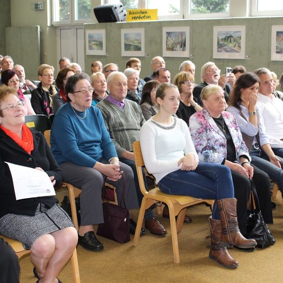 Ein Raum mit einer Gruppe von Menschen, die in Reihen von Stühlen sitzen, möglicherweise bei einer Besprechung. Ein Schild an der Wand sagt Raucher verboten.