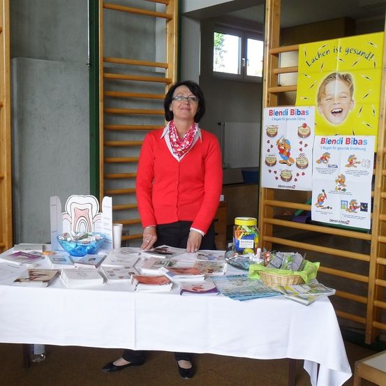 Eine Frau in einem roten Pullover steht hinter einem Tisch, der mit Büchern, Broschüren und einem Glas mit Süßigkeiten bedeckt ist. Der Tisch steht in einem Raum mit gelben Holzstangen und einem Poster mit einem lächelnden Kind.