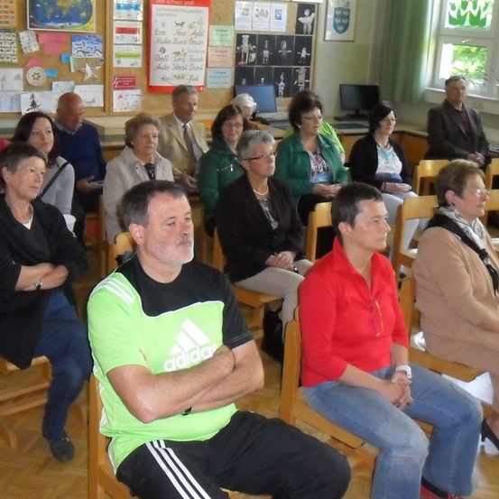 Eine Gruppe von Erwachsenen sitzt in Stühlen in einem Raum mit Pinnwänden und Fenstern.