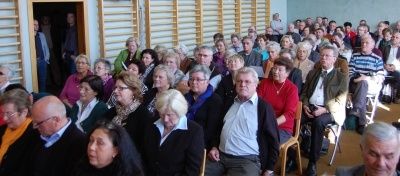 Eine große Menschenmenge, überwiegend ältere Erwachsene, sitzt in Stühlen in einem Saal, möglicherweise während einer Präsentation. Der Raum hat Holzwände und eine Tür im Hintergrund.