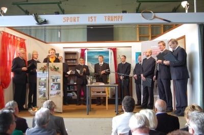 Eine Gruppe von Männern in Anzügen und Krawatten steht auf der Bühne mit einem Podium, Mikrofon und Tisch. Sie halten wahrscheinlich eine Rede oder Präsentation. Hinter ihnen ist eine Wand mit einem Gemälde, einem Regal mit Büchern und einem Vorhang. Das Publikum sitzt vor der Bühne.