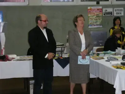 Ein älterer Mann und eine ältere Frau stehen zusammen in einem Raum mit Tischen, einer hält eine Mappe. Hinter ihnen sitzen und stehen andere Personen an den Tischen.