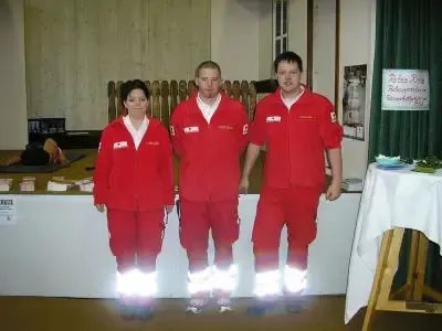 Drei Personen in roten Overalls mit reflektierenden Streifen stehen vor einem weißen Tisch mit weißer Tischdecke. Die Person links ist eine Frau, die beiden anderen sind Männer.