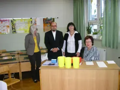 Vier Personen stehen in einem Klassenzimmer. Drei von ihnen stehen hinter einem Schreibtisch und eine sitzt in einem Stuhl. Ein Heizkörper ist an der Wand und ein Fenster mit grünen Vorhängen befindet sich rechts.