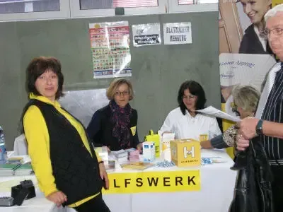 Fünf Personen stehen um einen Tisch mit einem gelben Schild, auf dem 'LFSWERK' steht. Eine Person hält ein Papier. Eine Frau in einer gelben und schwarzen Jacke steht links.