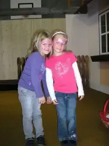 Zwei junge Mädchen stehen Seite an Seite in einem Raum. Das Mädchen links trägt ein lila Langarmshirt und Jeans. Das Mädchen rechts trägt ein rosa Shirt und Jeans. Beide haben ihre Hände vor sich verschränkt.