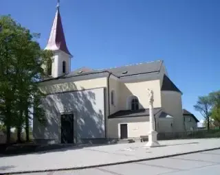 Eine weiße Kirche mit einem Turm, einer Statue vor dem Eingang und Bäumen an den Seiten unter einem klaren blauen Himmel.