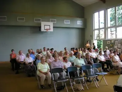 Eine große Gruppe älterer Menschen sitzt in einem Raum mit Basketballkorb an der Wand in Stühlen.