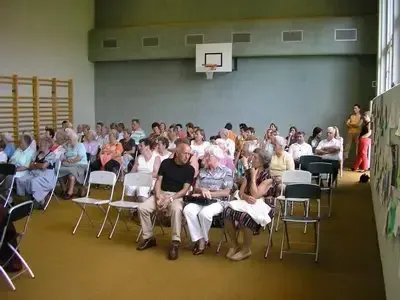 Eine große Gruppe älterer Menschen sitzt auf Stühlen in einem Saal mit einem Basketballkorb an der Wand und einer Wand mit Zeichnungen.