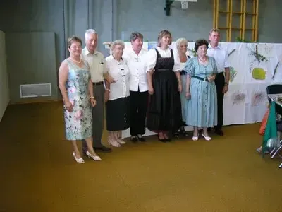 Eine Gruppe älterer Menschen posiert für ein Foto in einem Raum mit einem weißen Hintergrund, einem Holzstuhl und einer gelben Struktur im Hintergrund.