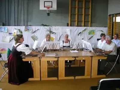 Eine Gruppe von Menschen in traditioneller Kleidung sitzt um einen Tisch in einem Raum mit Musikinstrumenten und einer Rückseite mit verschiedenen Zeichnungen.