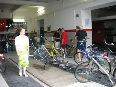 Mehrere Fahrräder sind in einer Garage geparkt. Eine erwachsene Frau steht neben einem umgefallenen Fahrrad, und mehrere erwachsene Männer stehen im Hintergrund.