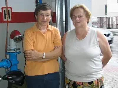 Zwei Frauen stehen vor einer Feuerwache, eine in einem weißen Tanktop und die andere in einem orangefarbenen Shirt. Ein blauer Feuerhydrant ist hinter ihnen sichtbar.