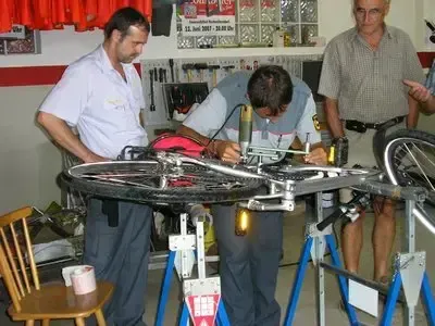 Drei Männer sind in einer Fahrradwerkstatt. Ein Mann arbeitet an einem Fahrradreifen, während die anderen beiden zusehen.