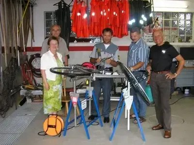 Fünf Personen stehen um einen Fahrradreparaturstand in einer Werkstatt herum. Drei Männer arbeiten am Fahrrad, während zwei Frauen zusehen.