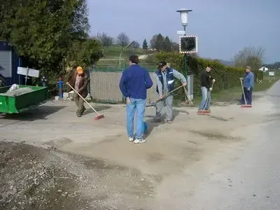 Sechs Männer fegen mit Besen eine Schotterstraße, einer mit einem grünen Anhänger in der Nähe.