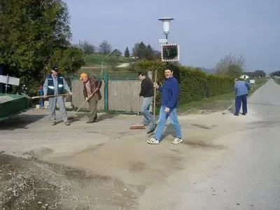 Fünf Männer sind auf einer Straße und benutzen Besen und Schaufeln, um die Straße zu reinigen. Ein Mann steht hinter einem Tor, und eine Ampel befindet sich auf der Straße.