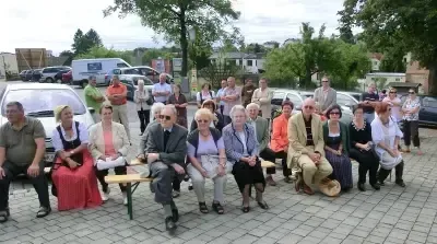Eine Gruppe älterer Menschen sitzt auf Bänken im Freien. Einige tragen Sonnenbrillen und Hüte. Autos, Bäume und Häuser sind im Hintergrund.