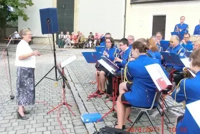 Eine Frau mit Brille spricht in ein Mikrofon, während eine Gruppe von Menschen in blauen Uniformen verschiedene Musikinstrumente auf einem gepflasterten Weg spielt.