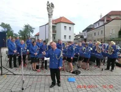 Eine Brassband steht in einer Reihe auf einem gepflasterten Platz vor einem Gebäude. Die Band besteht aus einem Dirigenten, Posaunen, Trompeten und Tuben. Datum und Uhrzeit sind unten rechts angegeben.