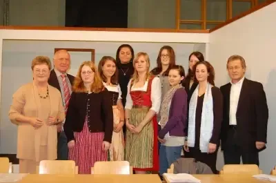 Eine Gruppe von Menschen steht in einem Raum und posiert möglicherweise für ein Foto. Sie tragen formelle Kleidung, und einige von ihnen tragen Brillen. Eine Frau trägt eine Halskette. Vor ihnen stehen einige Tische. Dahinter befindet sich eine weiße Wand, und einige Lampen sind an der Decke montiert.