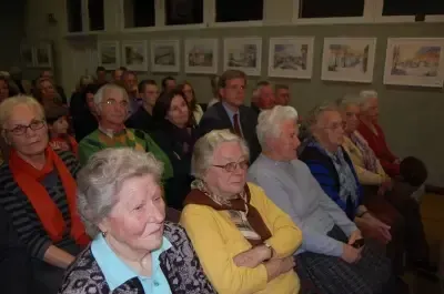 Eine Gruppe älterer Menschen sitzt in Stühlen in einem Raum mit gerahmten Bildern an der Wand.