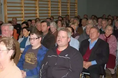 Eine große Menschenmenge, bestehend aus Männern und Frauen, sitzt in Stühlen in einem Raum. Einige tragen Brillen, während andere Mützen haben. Der Raum hat Holzwände und einen Holzboden.