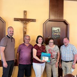 Fünf Menschen stehen lächelnd für ein Foto vor einer Kirchenwand. Eine Frau hält ein Buch mit dem Titel 'In Vielfalt'. Dahinter sind ein Kruzifix und ein gerahmtes Bild an der Wand angebracht.