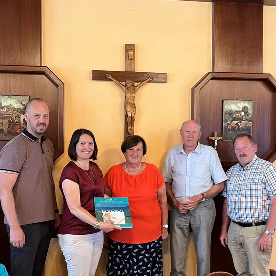 Fünf Erwachsene stehen vor einer Wand mit einem großen Kruzifix und gerahmten Gemälden. Die Frau in der Mitte hält ein Buch.